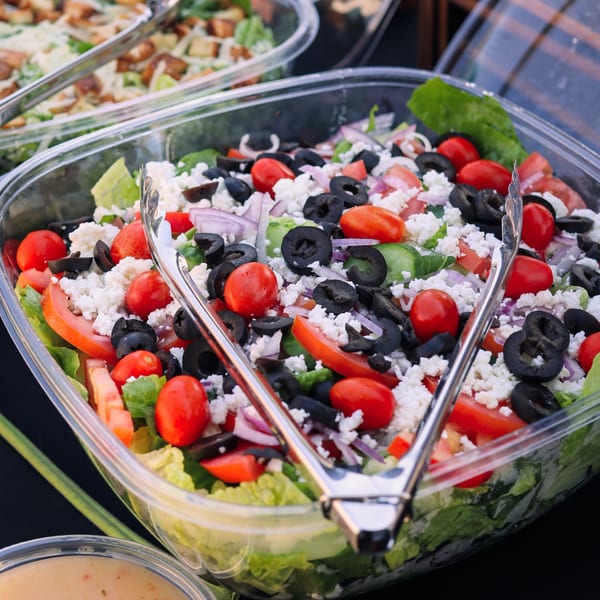 A large clear container of Greek salad with tomatoes, olives, feta, and red onion, with tongs.