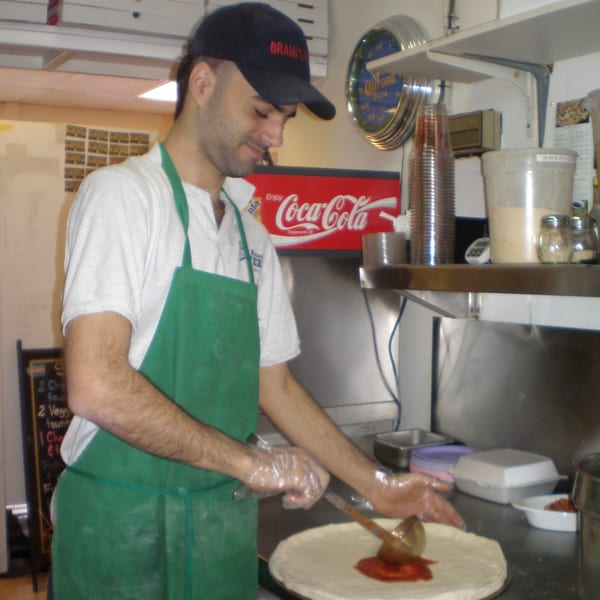 A pizza maker in a green apron and Brami's hat spreads sauce on a pizza crust.