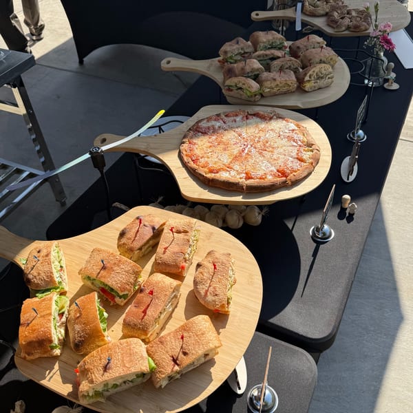 A black table with three wooden boards holding sandwiches, a cheese pizza, and bread knots.