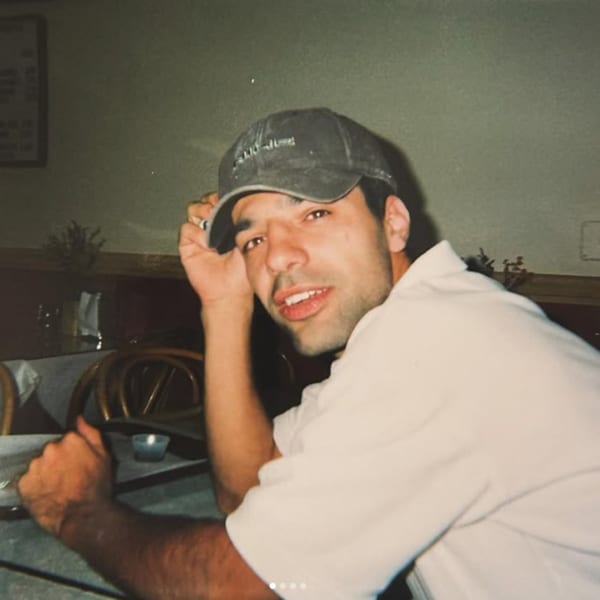 A man in a white shirt and dark baseball cap looks at the camera from a restaurant table.
