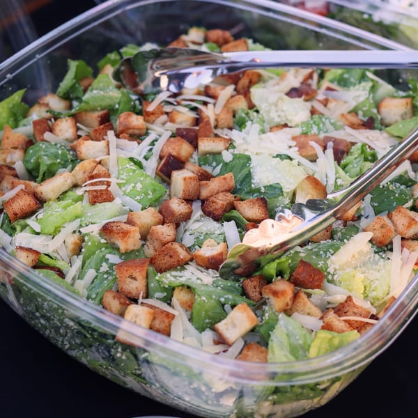 Caesar salad with romaine lettuce, croutons, and parmesan cheese in a clear plastic container with tongs.