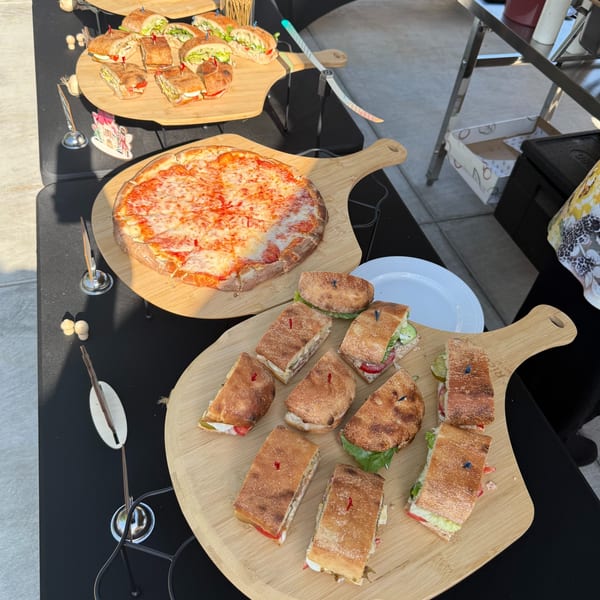 A buffet table with various sandwiches and a cheese pizza on wooden serving boards.