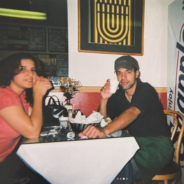 A man and woman sit at a table in a restaurant, the man holding a white cup.