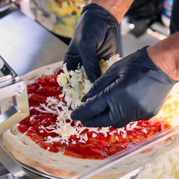 Hands in black gloves sprinkle shredded cheese onto a pizza with red sauce on a metal tray.