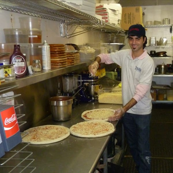 A pizza maker in a restaurant kitchen sprinkles cheese on pizzas, with ingredients and boxes around him.